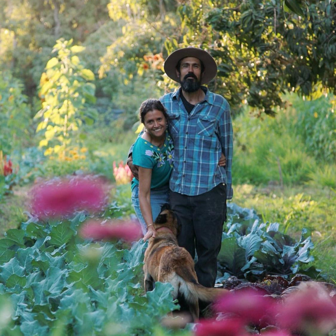 Two people and a dog in a garden with greenery and flowers.
